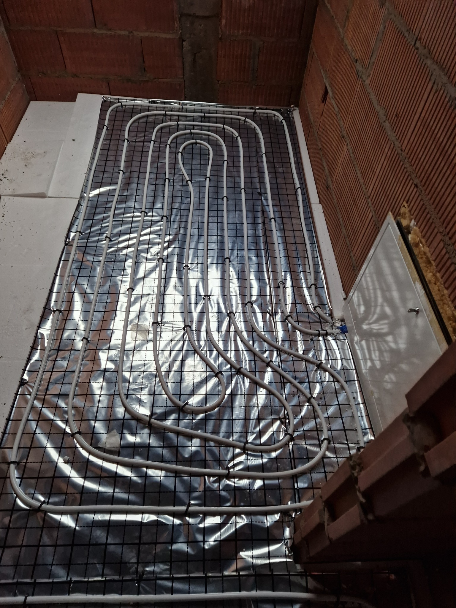 Białe rury ogrzewania podłogowego ułożone na srebrnej folii z czarną siatką, widoczne fragmenty muru z cegły i biały styropian.