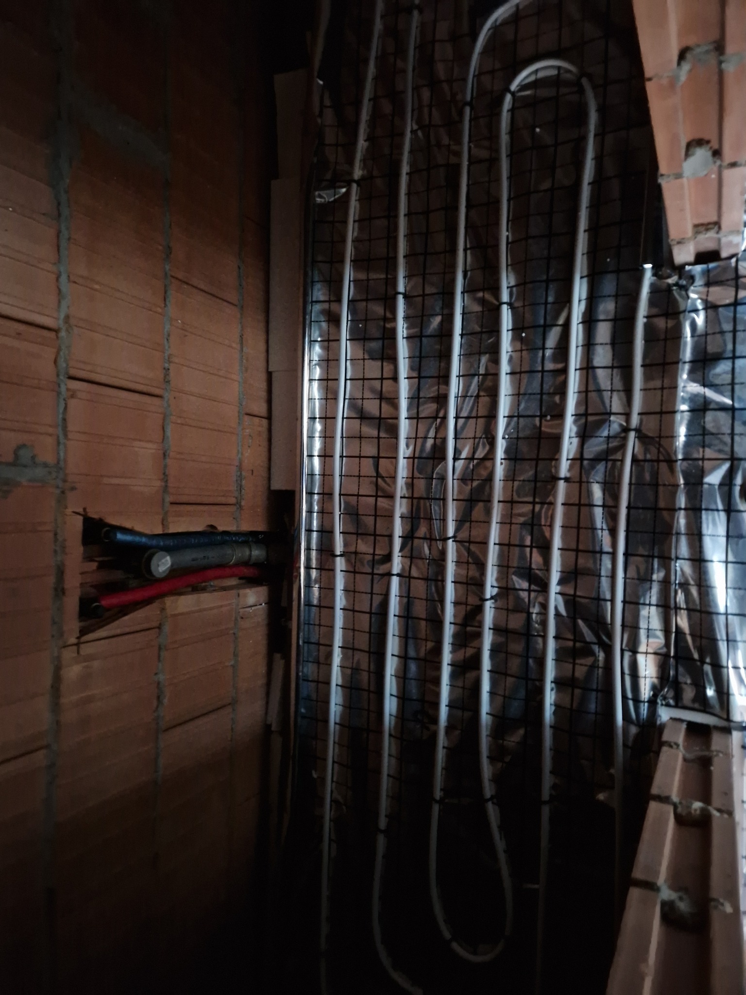 Montaż rur ogrzewania podłogowego na folii izolacyjnej z siatką, widoczne podłączenia rur w ścianie z cegły.