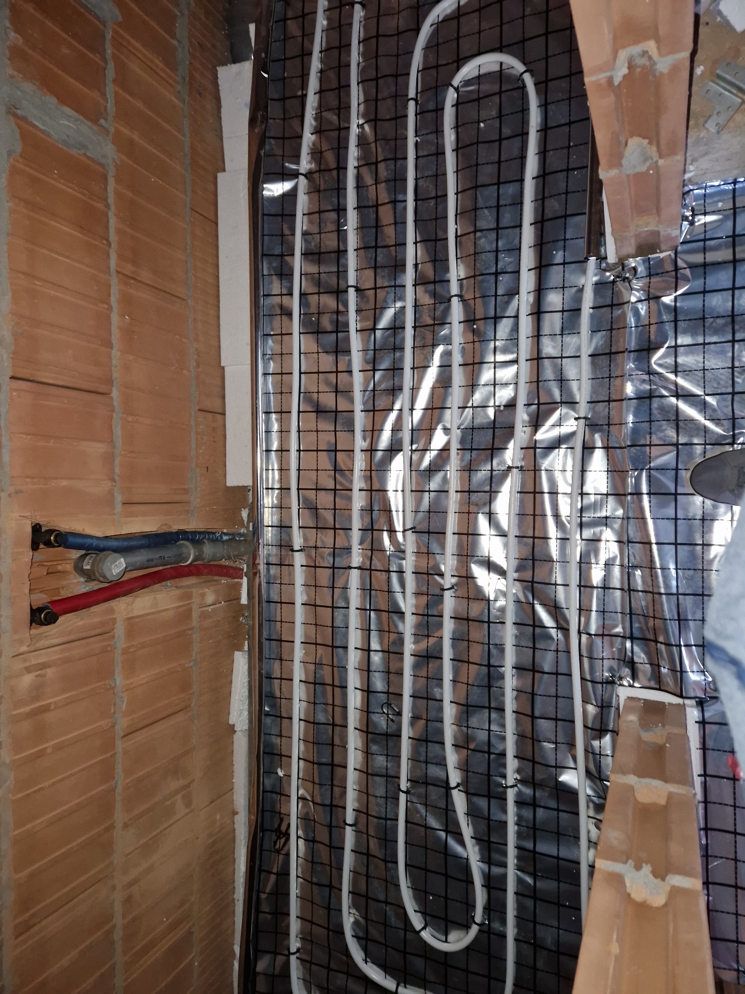 Montaż ogrzewania podłogowego: białe rurki grzewcze ułożone na srebrnej folii z czarną siatką, widoczne przyłącza rur do ściany z cegły.