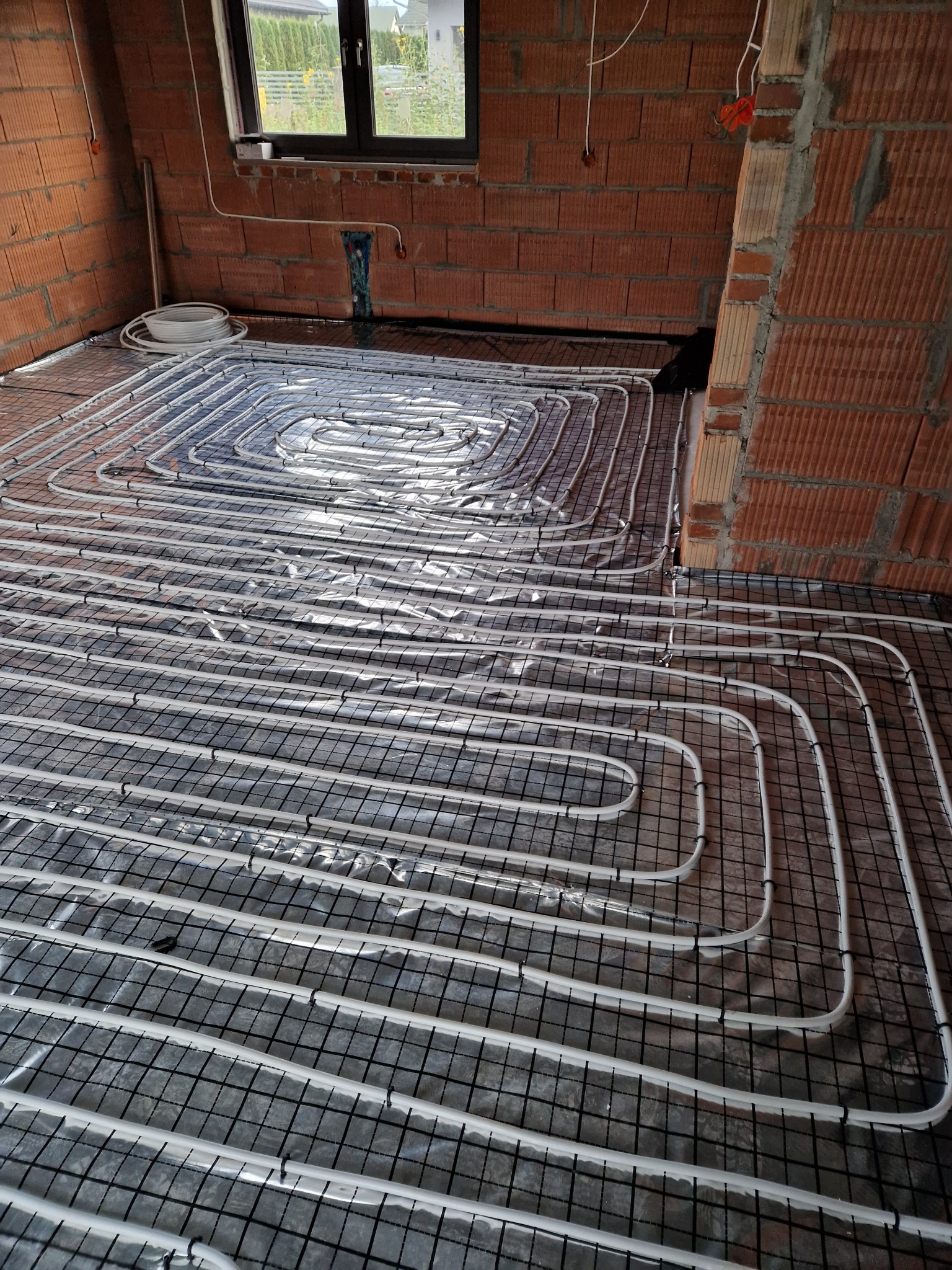Białe rurki ogrzewania podłogowego ułożone spiralnie na czarnej siatce w surowym wnętrzu z ceglanymi ścianami, widoczne okno i zwój rur.