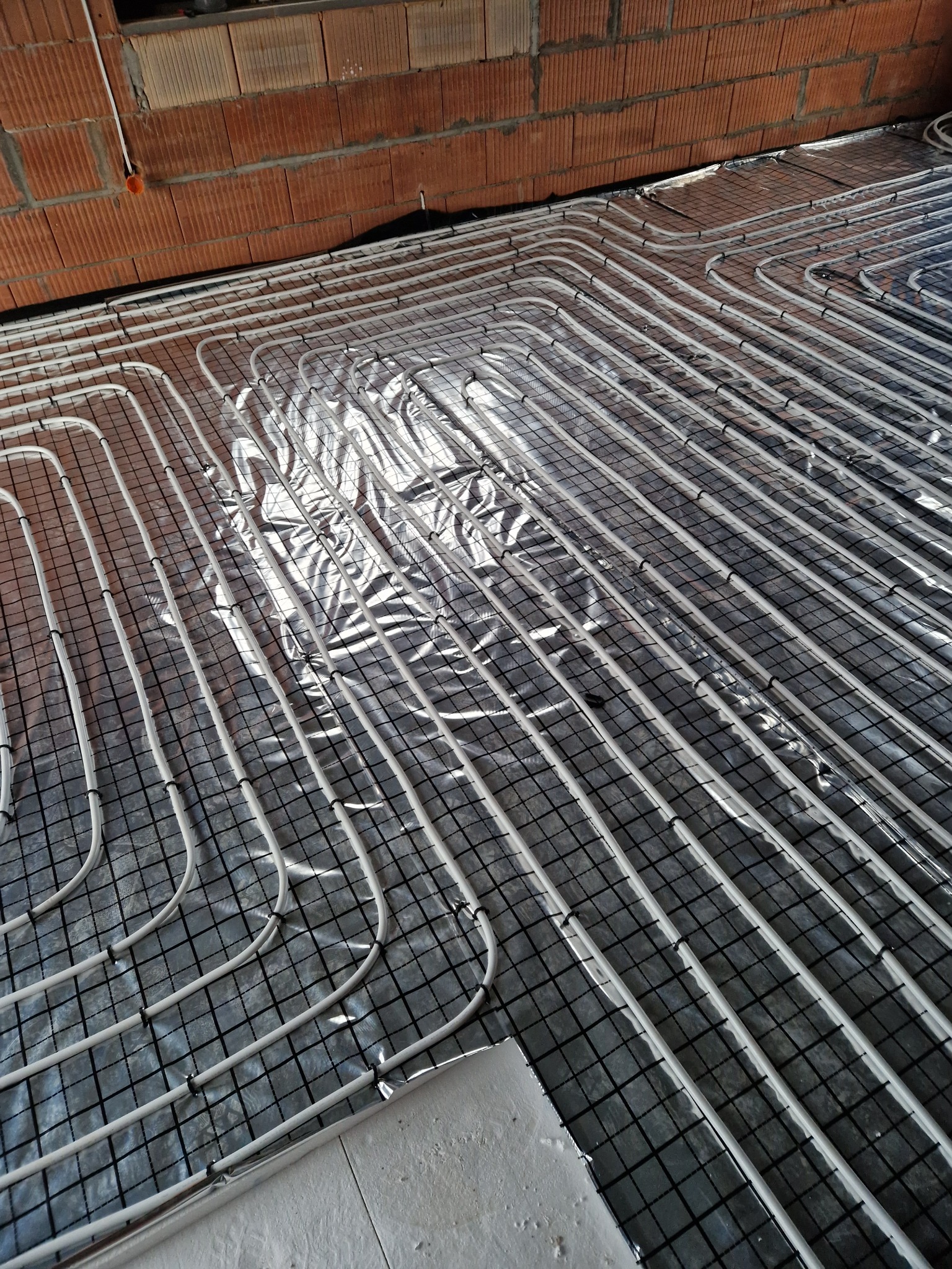Białe rury systemu ogrzewania podłogowego ułożone na folii izolacyjnej z czarną siatką, widoczne fragmenty ceglanej ściany i betonowej wylewki.
