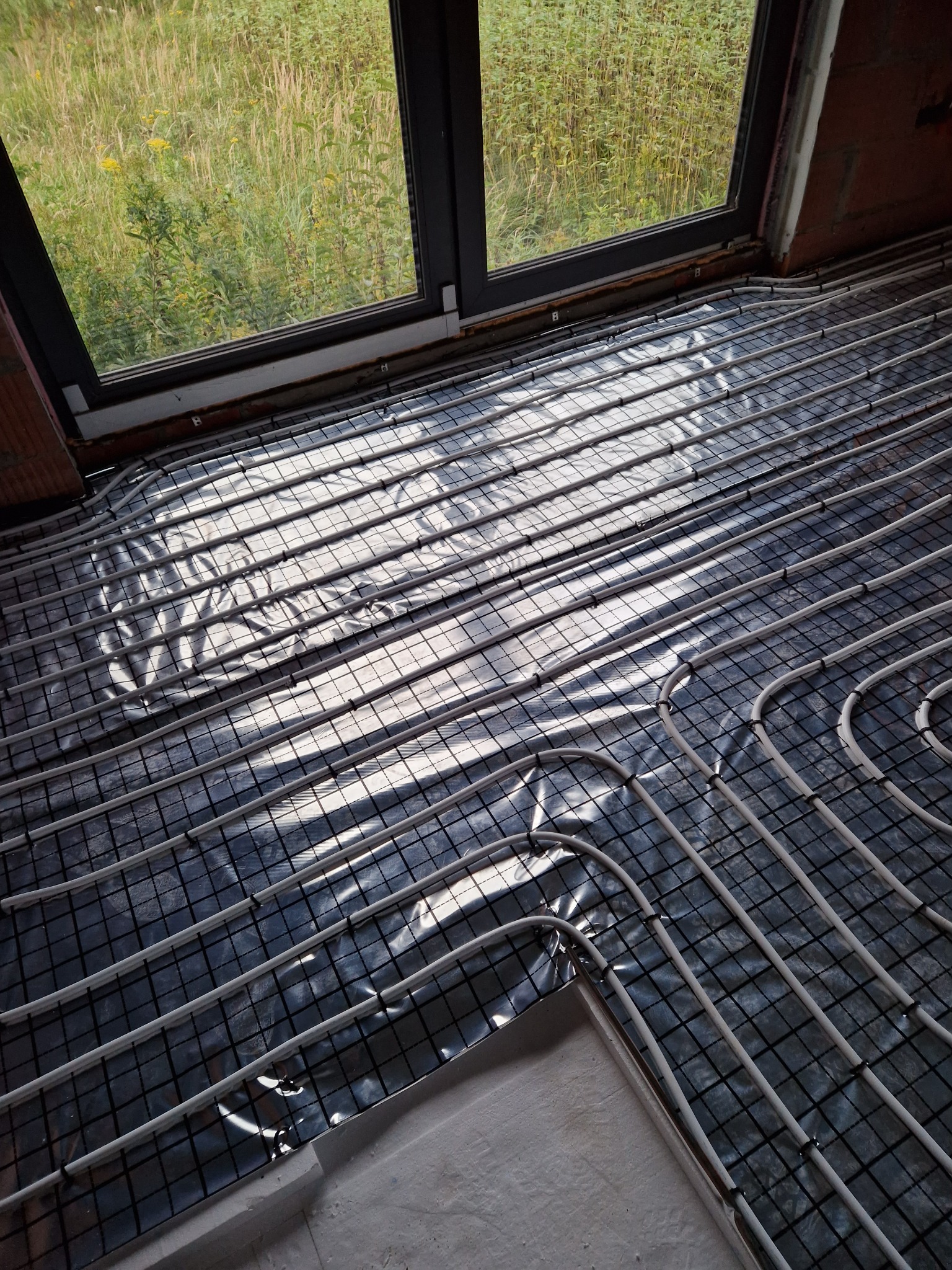 Białe rury ogrzewania podłogowego ułożone na czarnej folii z siatką, widok przez okno na zewnątrz budynku w trakcie montażu instalacji.