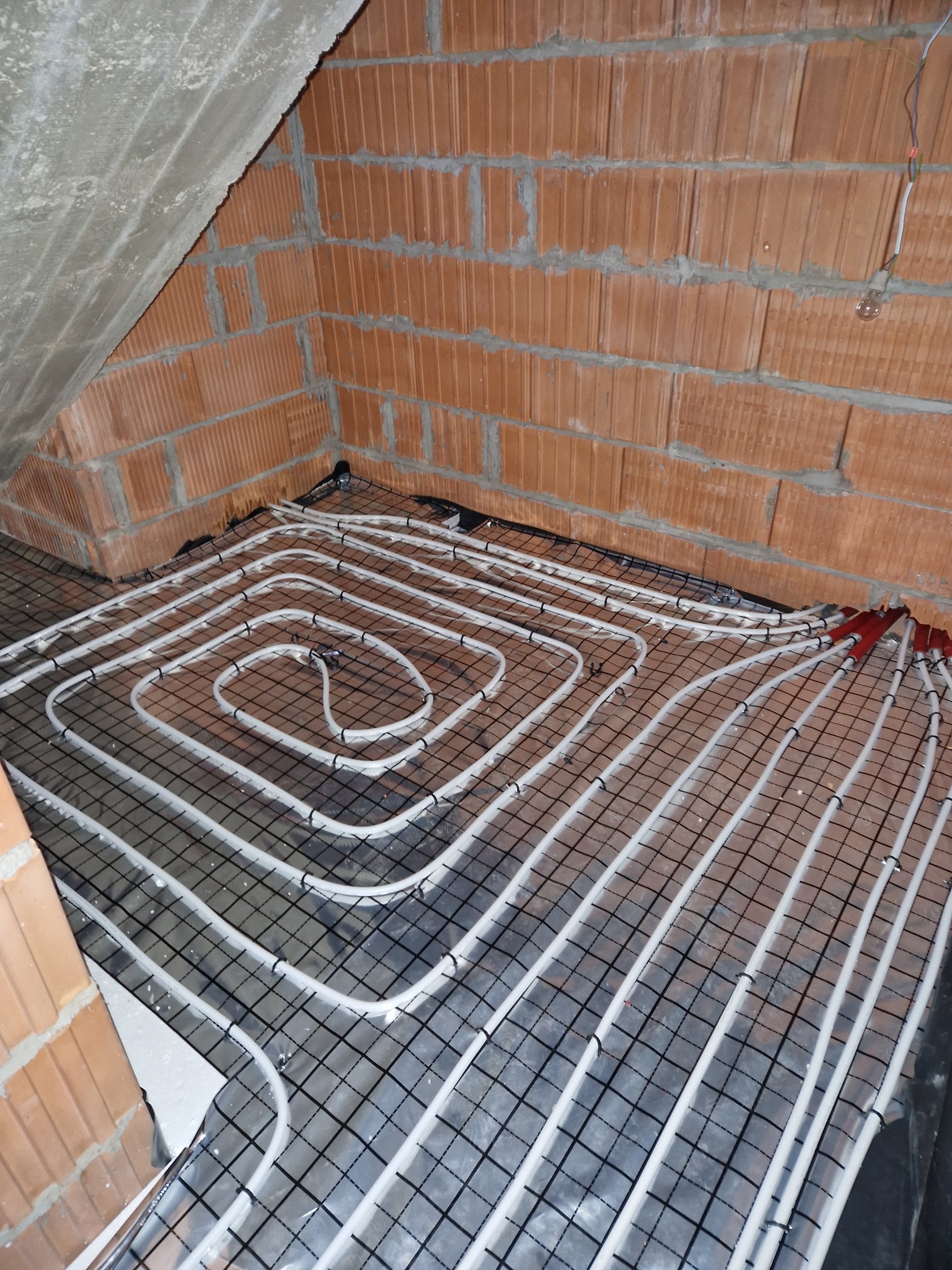 Układanie białych rur grzewczych na czarnej siatce podłogowej w surowym wnętrzu z ceglanymi ścianami i betonowym stropem.