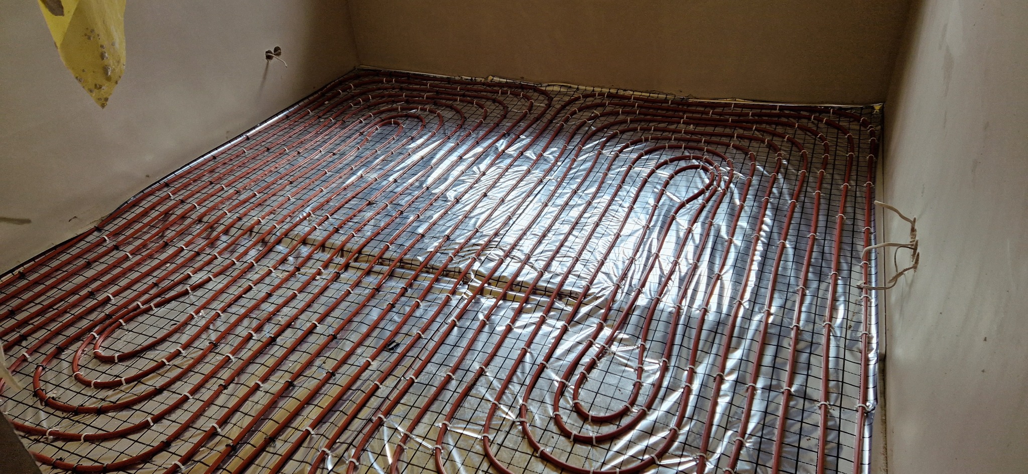 Rozłożona instalacja ogrzewania podłogowego z rurkami w kolorze ceglastym, zamocowanymi do siatki na folii izolacyjnej, widok z góry w surowym wnętrzu.