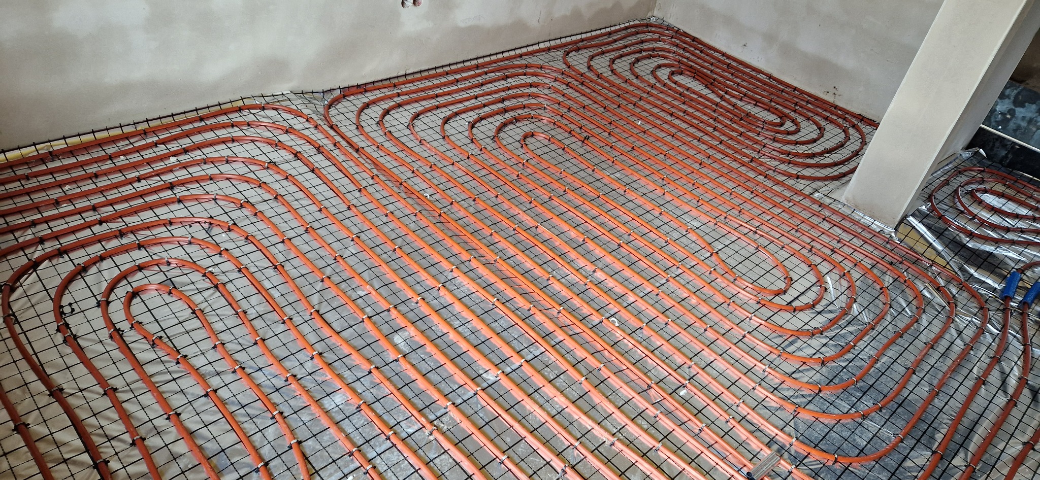 Pomarańczowe rury ogrzewania podłogowego ułożone na folii izolacyjnej i siatce montażowej, widok z góry na fragment instalacji w surowym stanie.