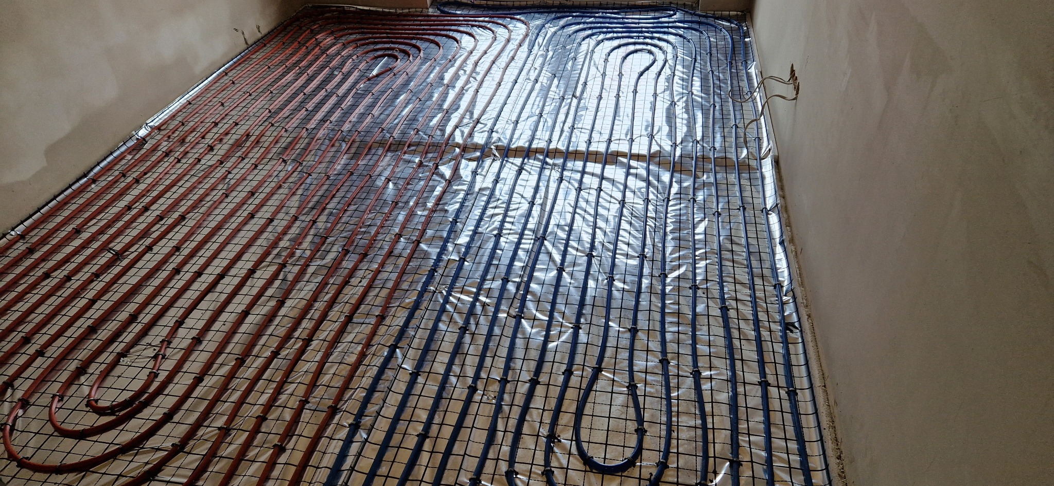Rozłożona instalacja ogrzewania podłogowego na folii izolacyjnej, z rurami w kolorze czerwonym i niebieskim, przymocowanymi do siatki, widoczne nieotynkowane ściany.