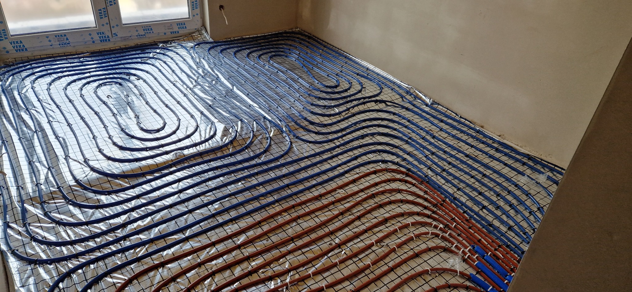 Rozłożone, niebieskie i czerwone rury ogrzewania podłogowego na folii izolacyjnej z siatką, widok z góry na wczesnym etapie instalacji w surowym wnętrzu.