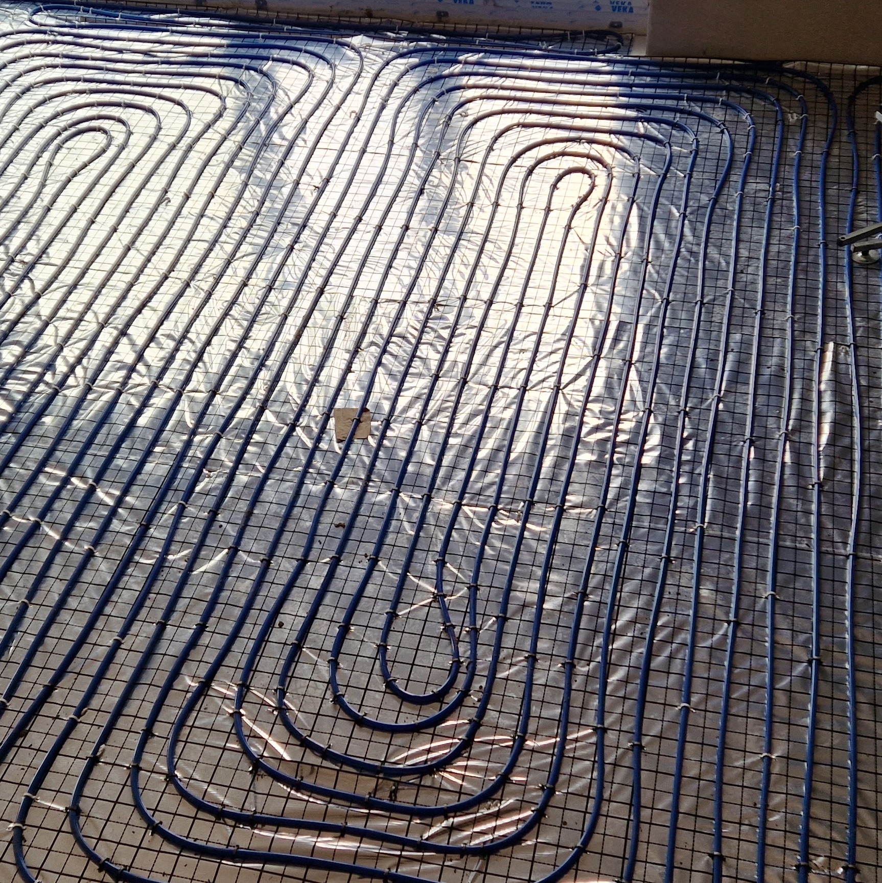Instalacja ogrzewania podłogowego z niebieskich rurek na folii izolacyjnej, przymocowanych do metalowej siatki, widoczne z góry w jasnym świetle.