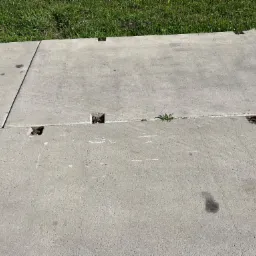 Szare płyty betonowe z widocznymi szczelinami i niewielkimi plamami, z trawą rosnącą w tle i w otworach montażowych.