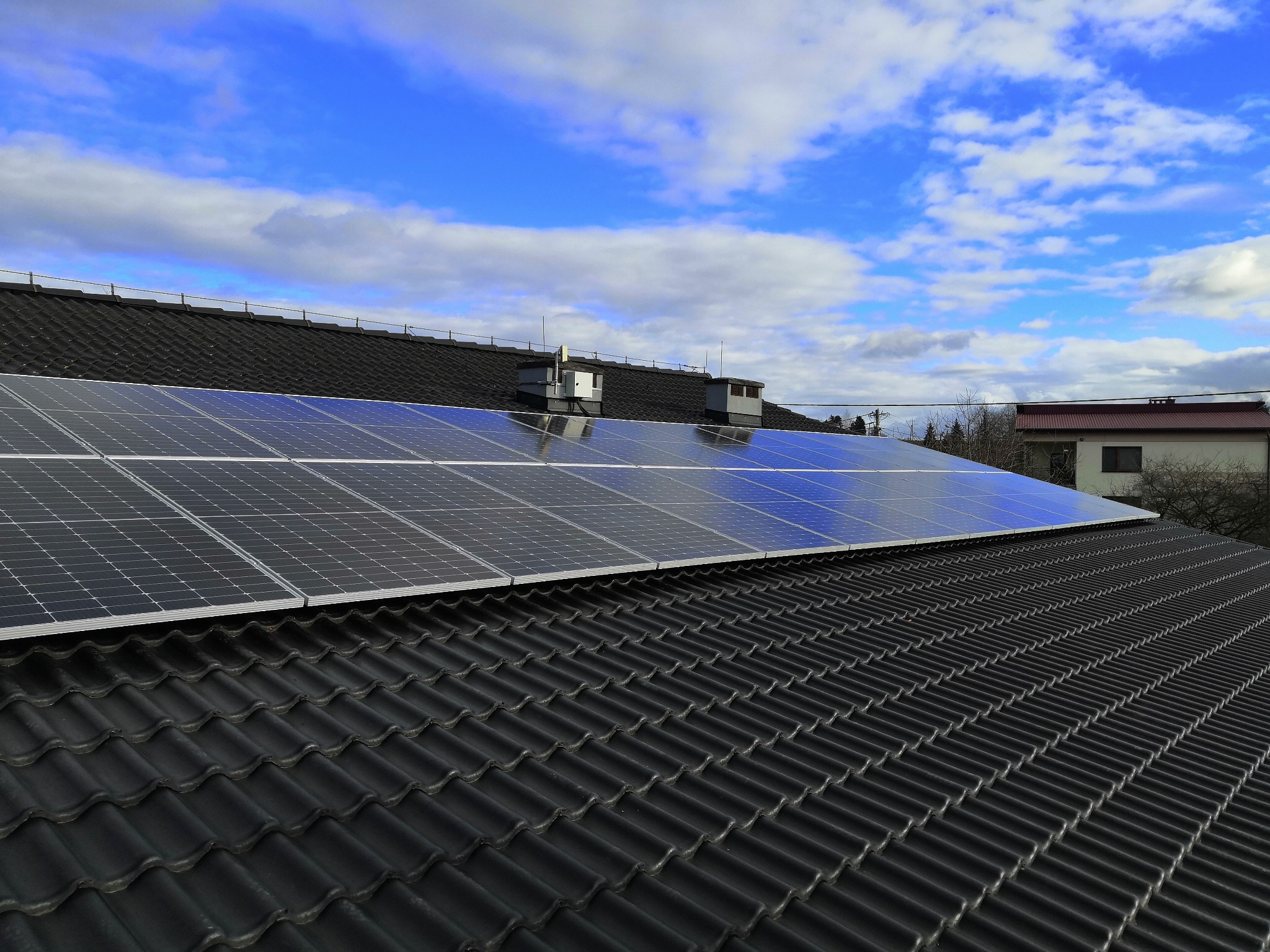 Panel słoneczny na ciemnym dachu z dachówki ceramicznej, odbijający błękitne niebo z białymi chmurami.