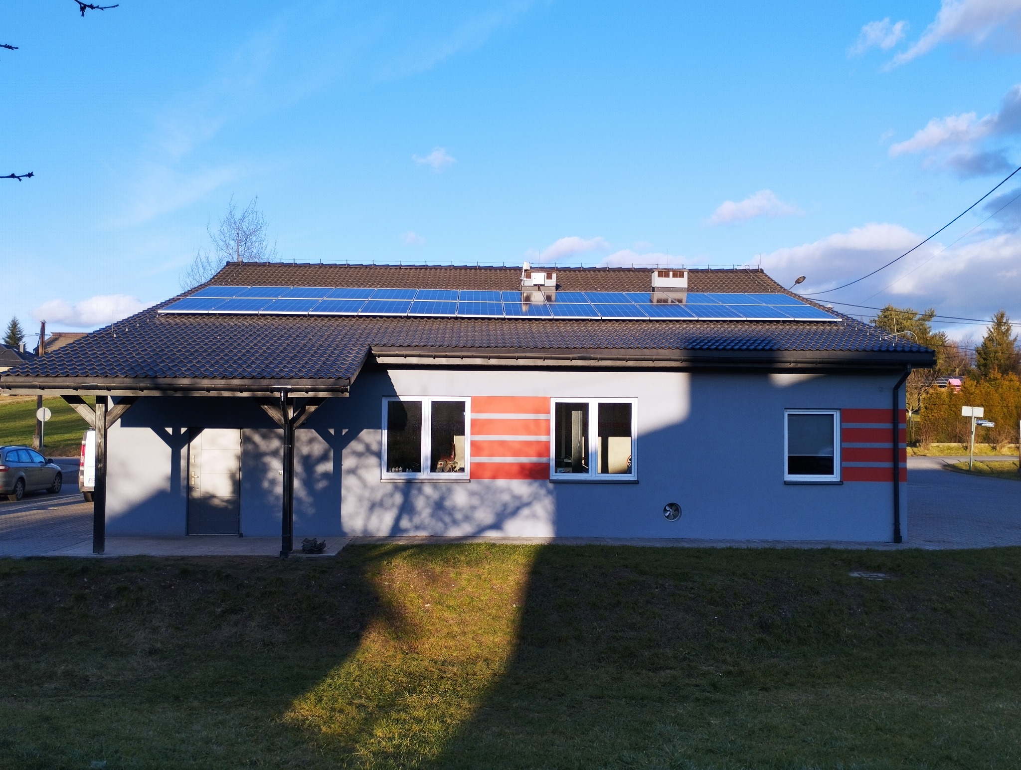 Niski budynek z szarą elewacją i panelami słonecznymi na dachu, widoczne trzy okna z czerwonymi pasami, trawnik przed budynkiem, błękitne niebo z chmurami.