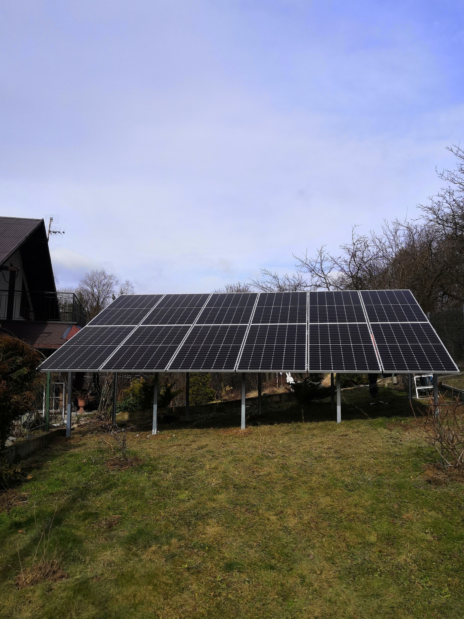 Panel słoneczny na metalowej konstrukcji w ogrodzie, widoczny dom z brązowym dachem w tle, pochmurne niebo.