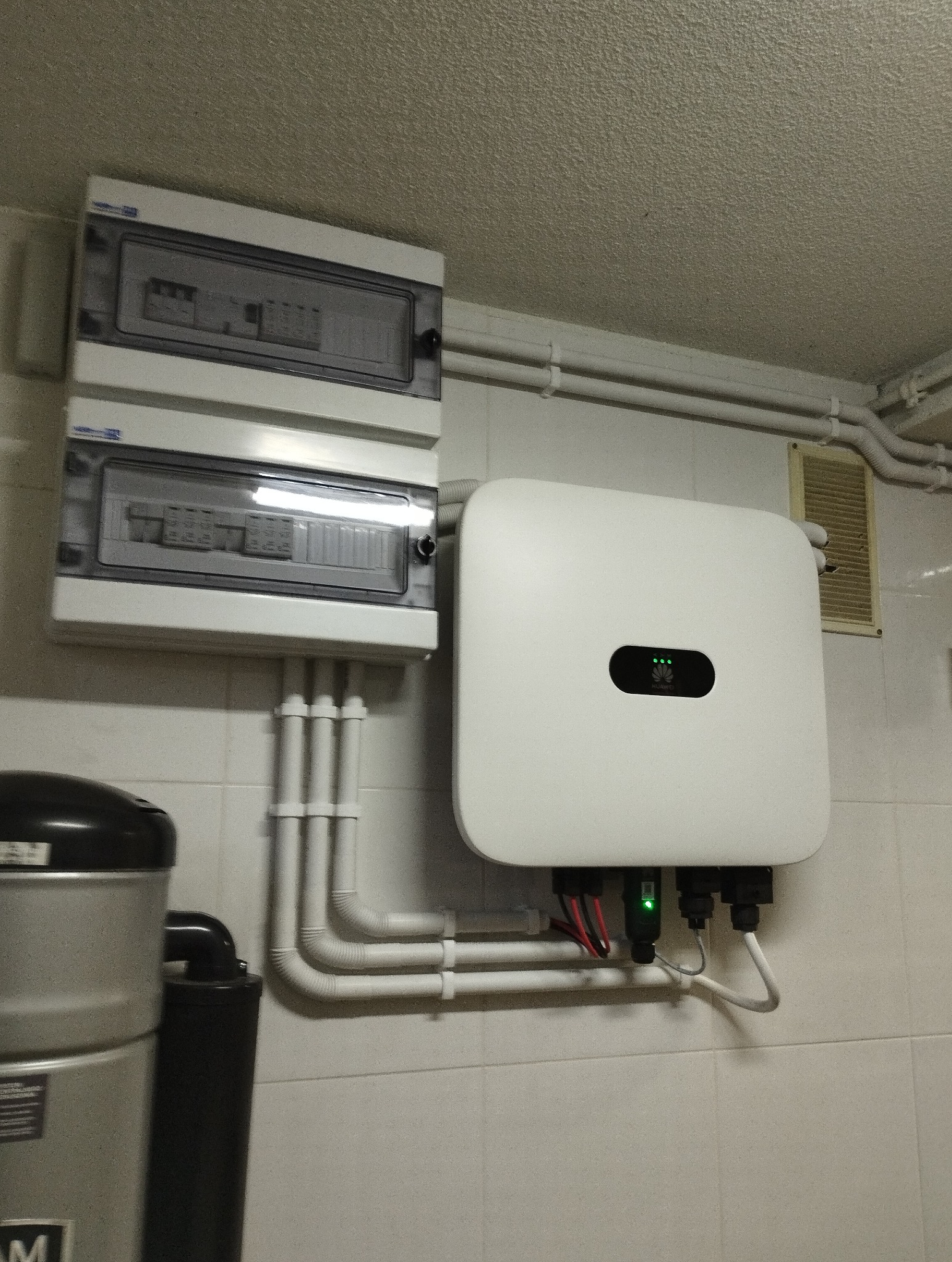 Biały falownik z trzema zielonymi diodami, podłączony przewodami do instalacji, obok skrzynka bezpiecznikowa na kafelkowanej ścianie w pomieszczeniu technicznym.