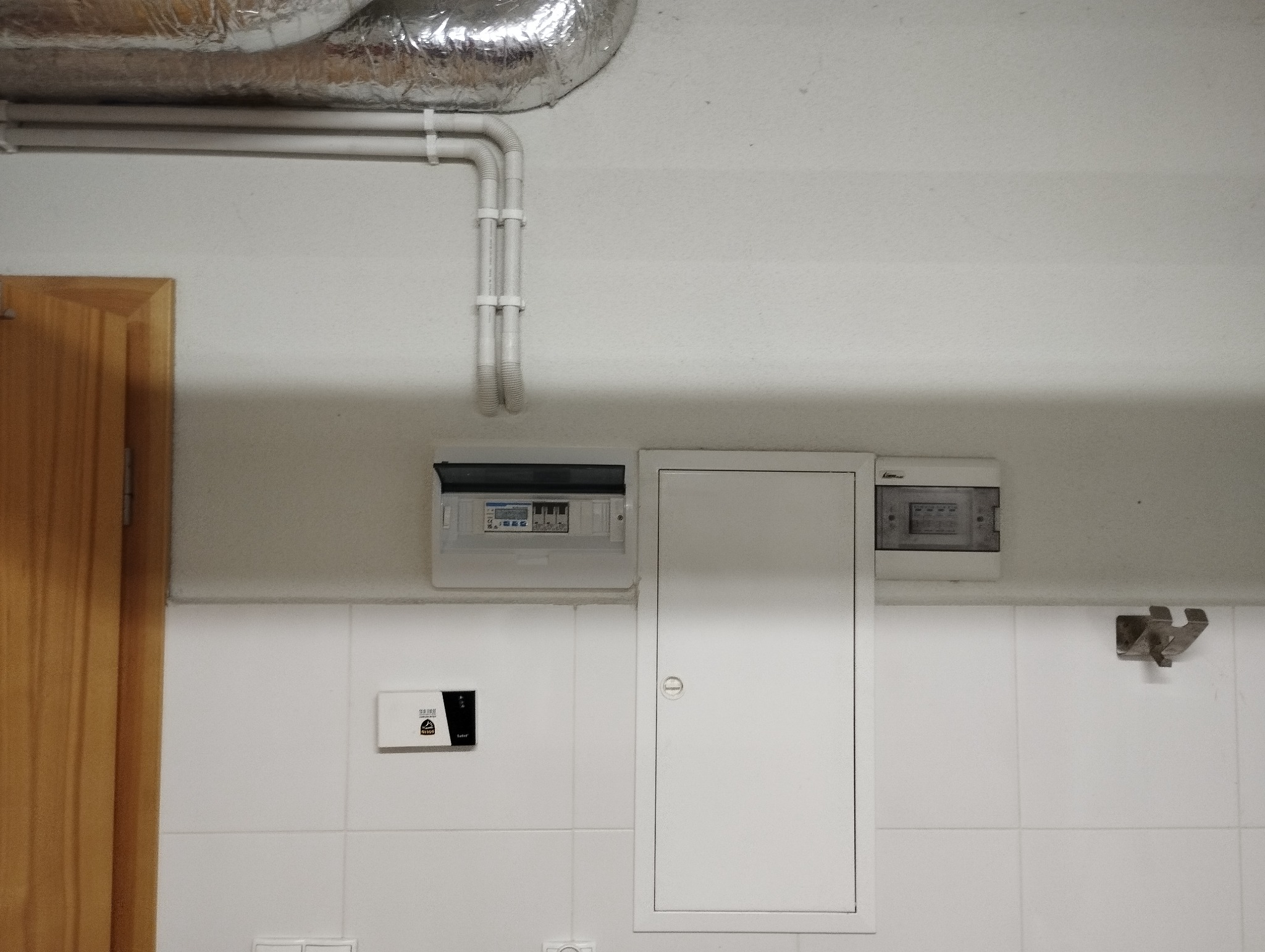 Rozdzielnia elektryczna z bezpiecznikami, rury instalacyjne pod sufitem, białe kafelki na ścianie i drewniane drzwi w tle.