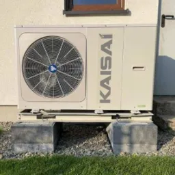 Zewnętrzna jednostka pompy ciepła Kaisai zamontowana na betonowych blokach przy ścianie budynku, widoczna trawa i kamyki.