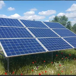 Instalacja fotowoltaiczna na metalowej konstrukcji, ustawiona na łące z polnymi kwiatami, w tle budynek gospodarczy i błękitne niebo z chmurami.