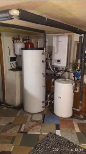 Instalacja grzewcza w piwnicy: Dwa białe zbiorniki akumulacyjne ciepła, wiszący bojler elektryczny i rury izolowane termicznie, widoczne na tle kafelkowej podłogi.