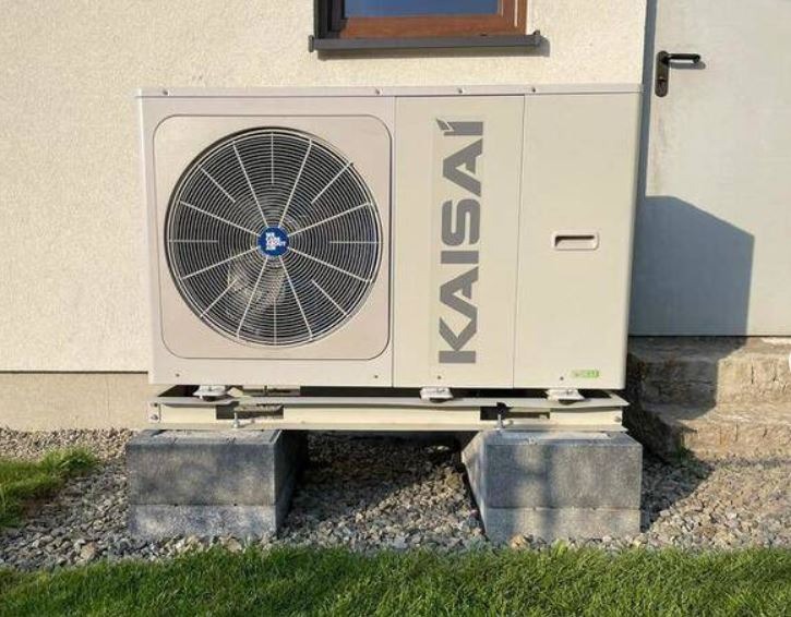 Zewnętrzna jednostka pompy ciepła Kaisai zamontowana na betonowych blokach przy ścianie budynku, widoczna trawa i kamyki.