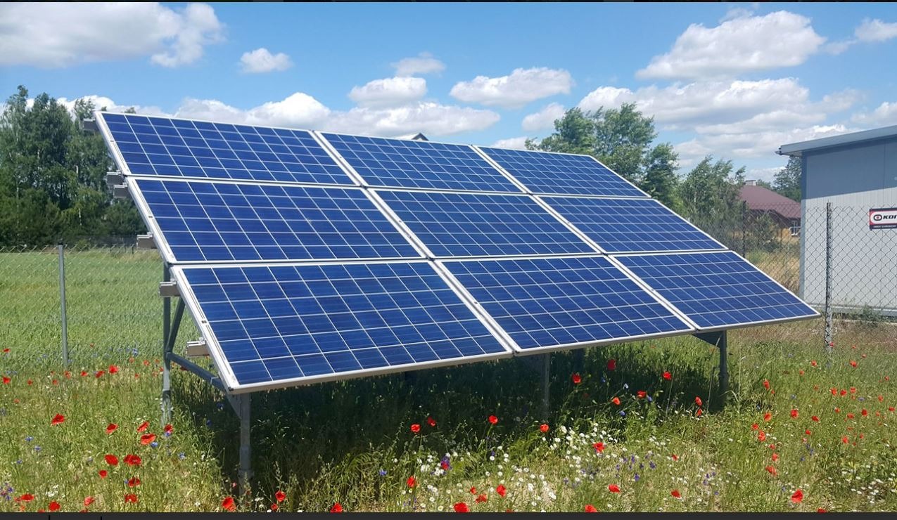 Instalacja fotowoltaiczna na metalowej konstrukcji, ustawiona na łące z polnymi kwiatami, w tle budynek gospodarczy i błękitne niebo z chmurami.