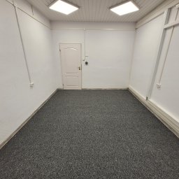 TM Projekt - Puste, odnowione pomieszczenie z szarym dywanem, białą teksturą ścian, białą stolarką drzwiową i dwoma kwadratowymi lampami sufitowymi.