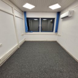 TM Projekt - Puste pomieszczenie po remoncie: szara wykładzina, białe ściany z widocznymi listwami przypodłogowymi, okno z niebieskimi zasłonami, klimatyzator na ścianie i kwadratowe lampy sufitowe.