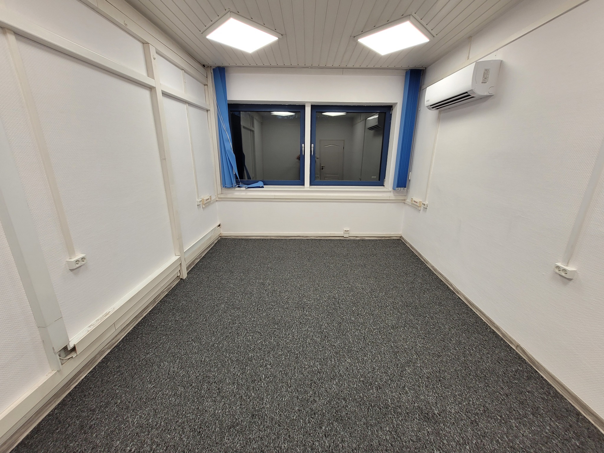 Puste pomieszczenie po remoncie: szara wykładzina, białe ściany z widocznymi listwami przypodłogowymi, okno z niebieskimi zasłonami, klimatyzator na ścianie i kwadratowe lampy sufitowe.