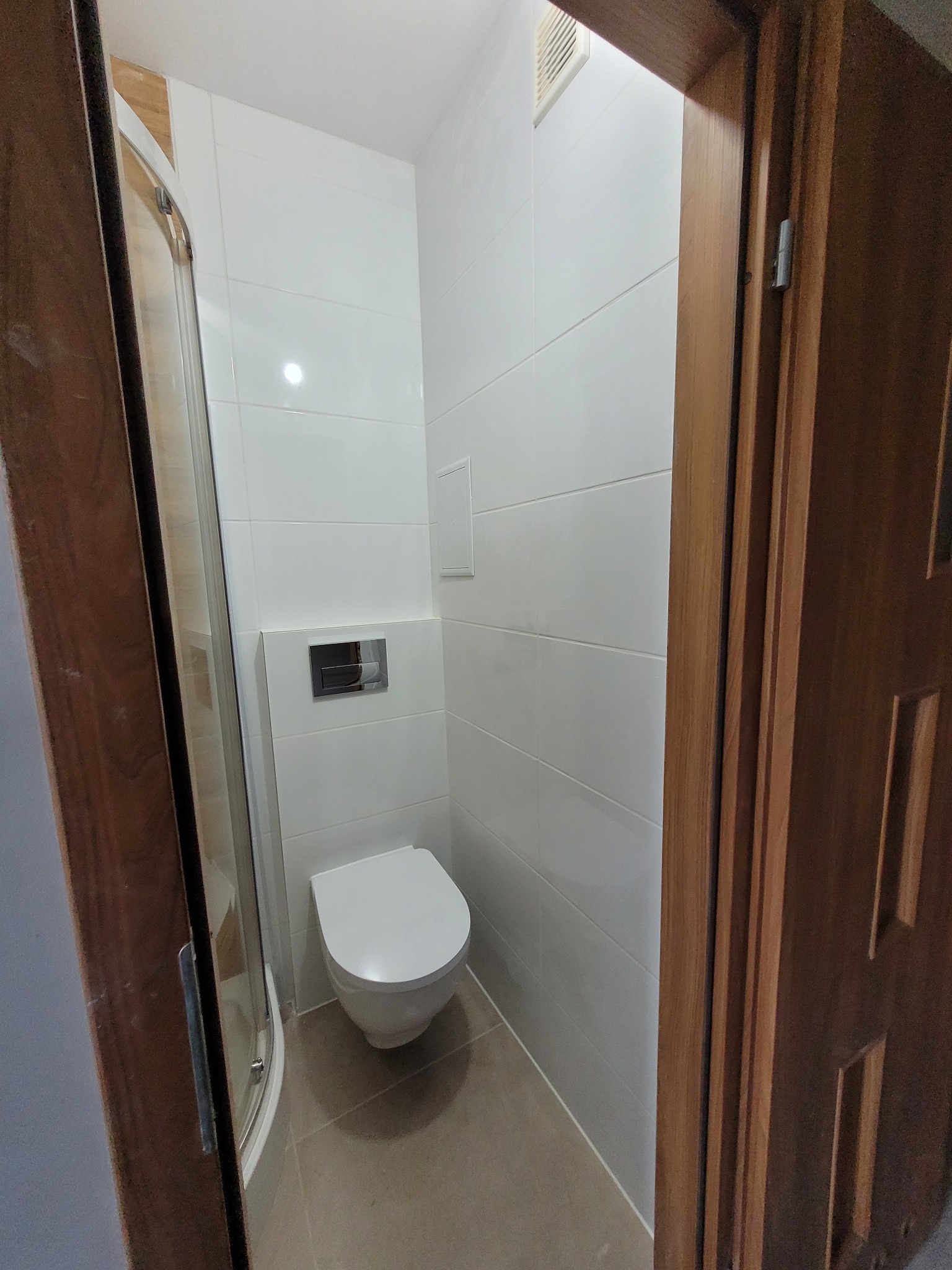 Wnętrze małej, wykończonej łazienki z białą toaletą wiszącą, jasnymi płytkami na ścianach i podłodze oraz częściowo widoczną kabiną prysznicową z zaokrąglonymi drzwiami, widzianej zza drewnianej...