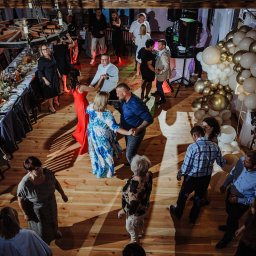 R&W Fotografia - Zabawa taneczna na sali z drewnianą podłogą, udekorowanej stołami i balonami w odcieniach bieli i złota, oświetlona ciepłym światłem.