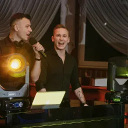 Dwóch uśmiechniętych prezenterów z mikrofonami na tle sceny z reflektorami i laptopem.