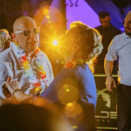 R&W Fotografia - Para tańczy na parkiecie podczas imprezy, mężczyzna w białej koszuli i okularach przeciwsłonecznych ma na szyi hawajski wieniec, w tle inni goście i oświetlenie sceniczne.