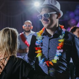 R&W Fotografia - Młody mężczyzna w kapeluszu i okularach z numerem 18, z hawajskim naszyjnikiem, uśmiecha się na tle sali balowej z balonami i innymi gośćmi.