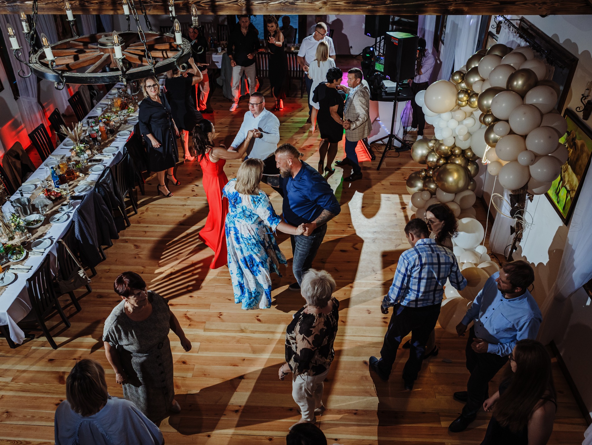 Zabawa taneczna na sali z drewnianą podłogą, udekorowanej stołami i balonami w odcieniach bieli i złota, oświetlona ciepłym światłem.