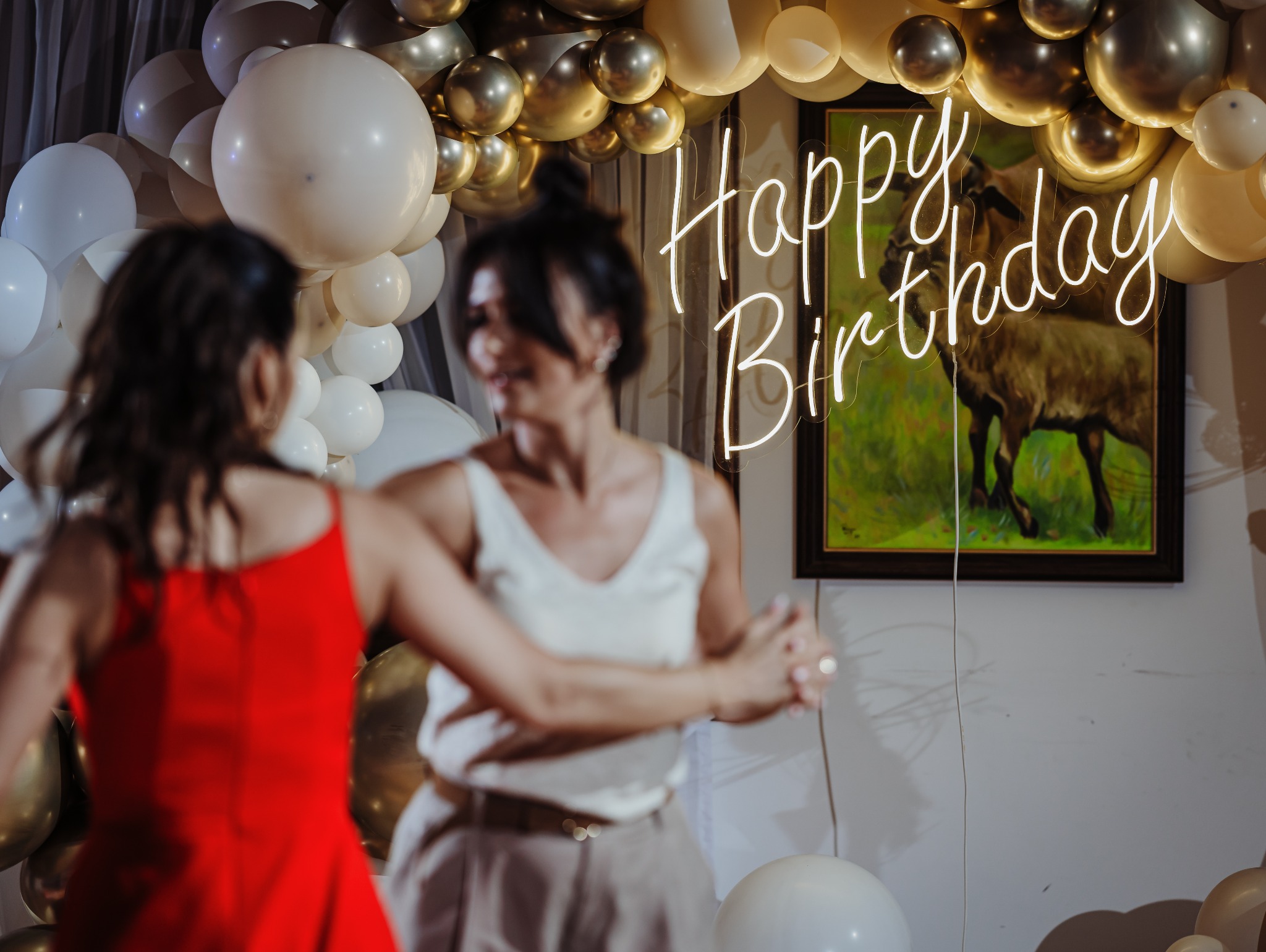Dwie kobiety w tańcu, dekoracja z białych i złotych balonów, neon 'Happy Birthday' i obraz z motywem zwierzęcym w tle.