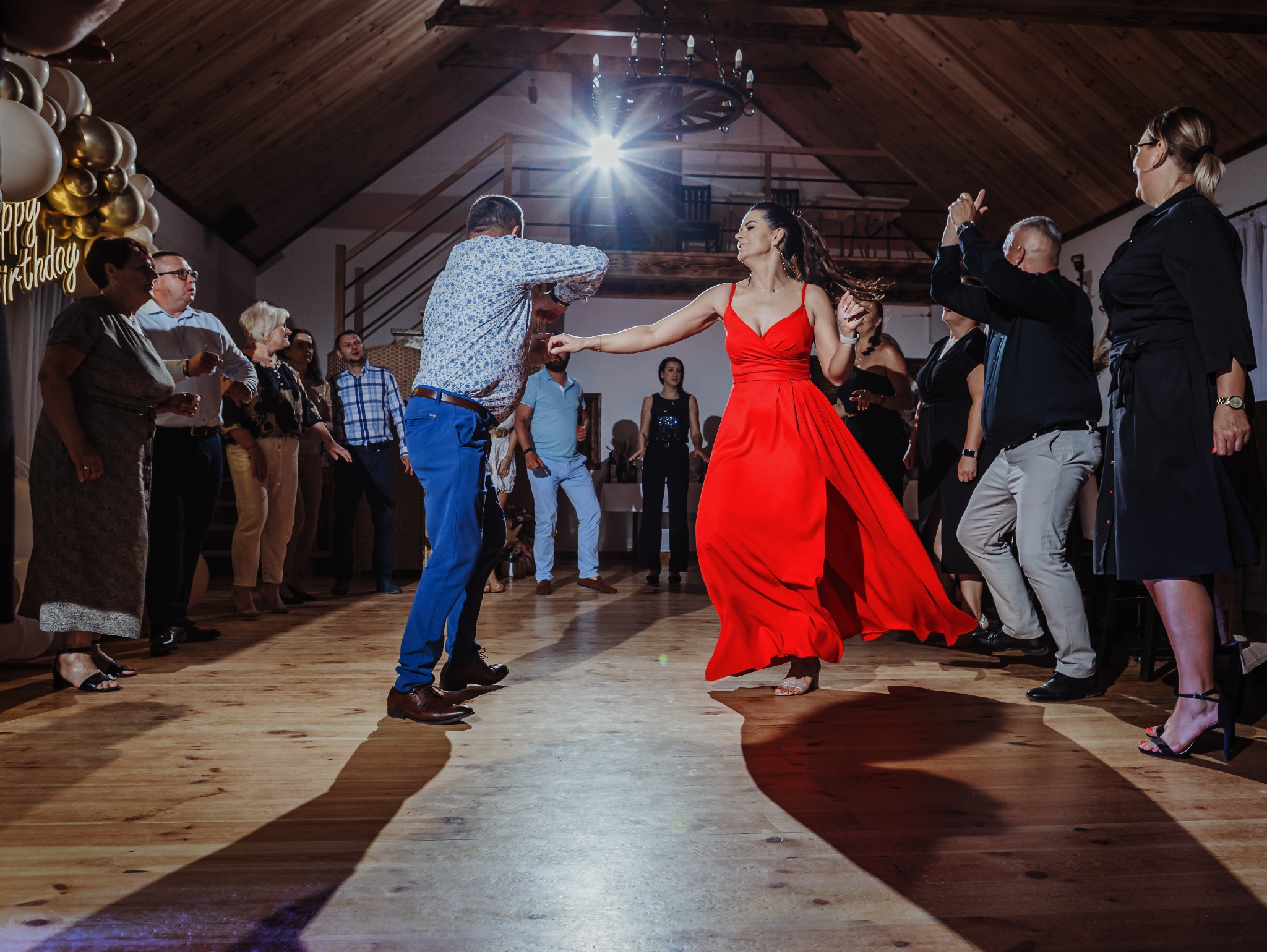 Uczestnicy tańczący na przyjęciu urodzinowym w sali z drewnianym parkietem i dekoracjami balonowymi, kobieta w czerwonej sukni w centrum kadru.