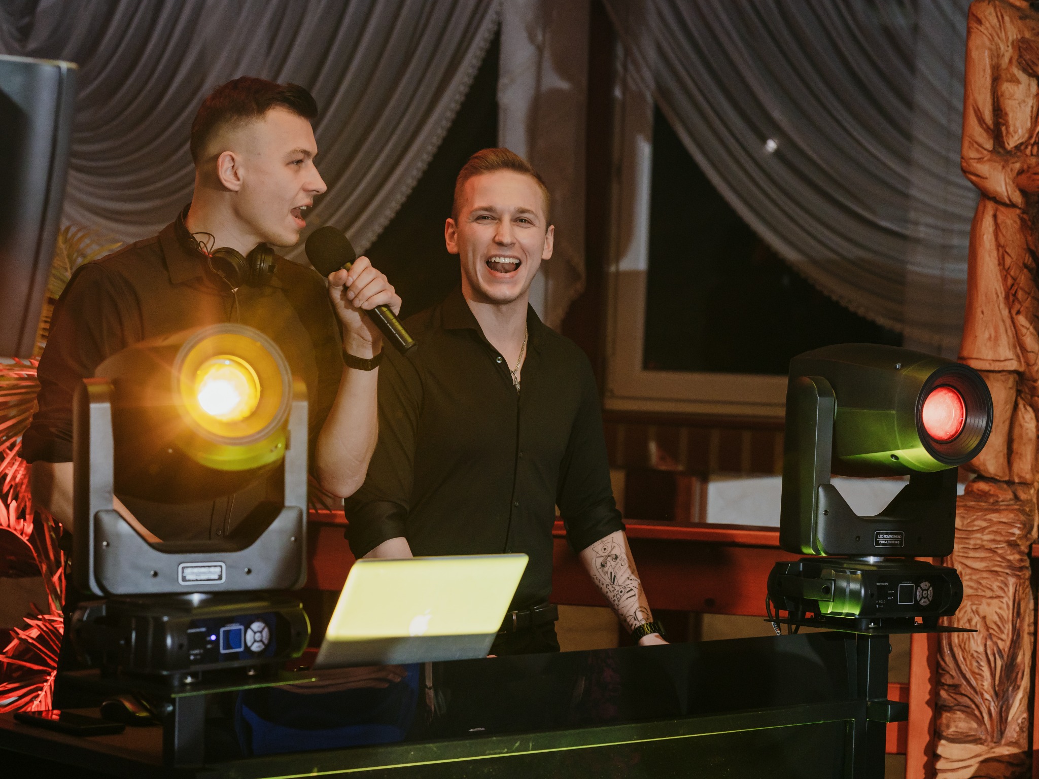 Dwóch uśmiechniętych prezenterów z mikrofonami na tle sceny z reflektorami i laptopem.