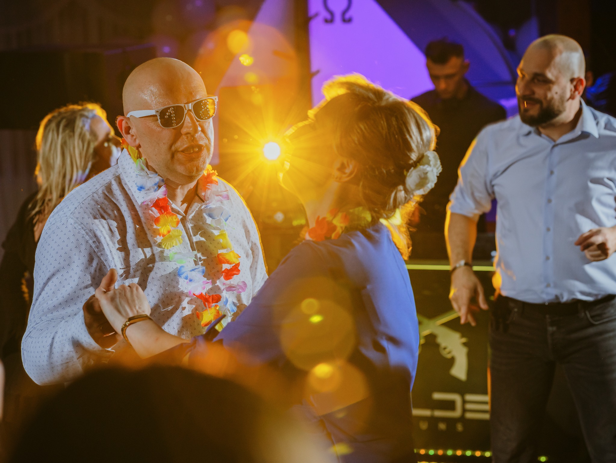 Para tańczy na parkiecie podczas imprezy, mężczyzna w białej koszuli i okularach przeciwsłonecznych ma na szyi hawajski wieniec, w tle inni goście i oświetlenie sceniczne.