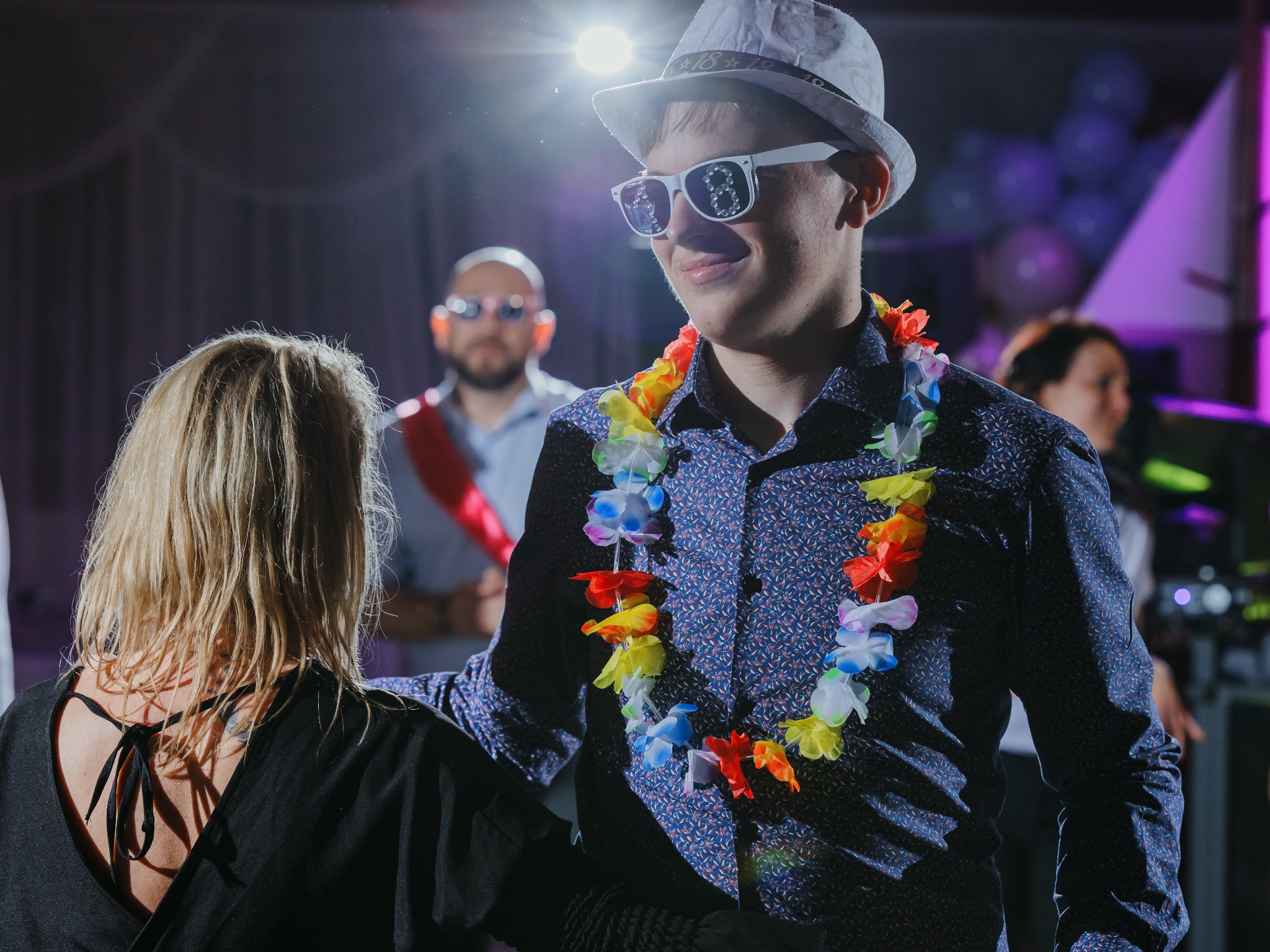 Młody mężczyzna w kapeluszu i okularach z numerem 18, z hawajskim naszyjnikiem, uśmiecha się na tle sali balowej z balonami i innymi gośćmi.