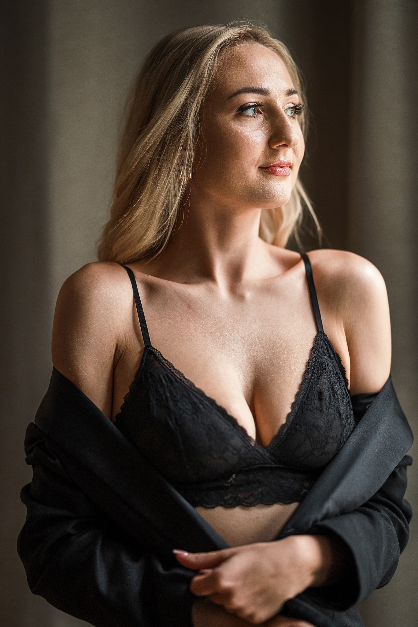 Portret kobiety o blond włosach, ubrana w koronkowy biustonosz i czarną marynarkę, spoglądająca w bok w miękkim świetle.