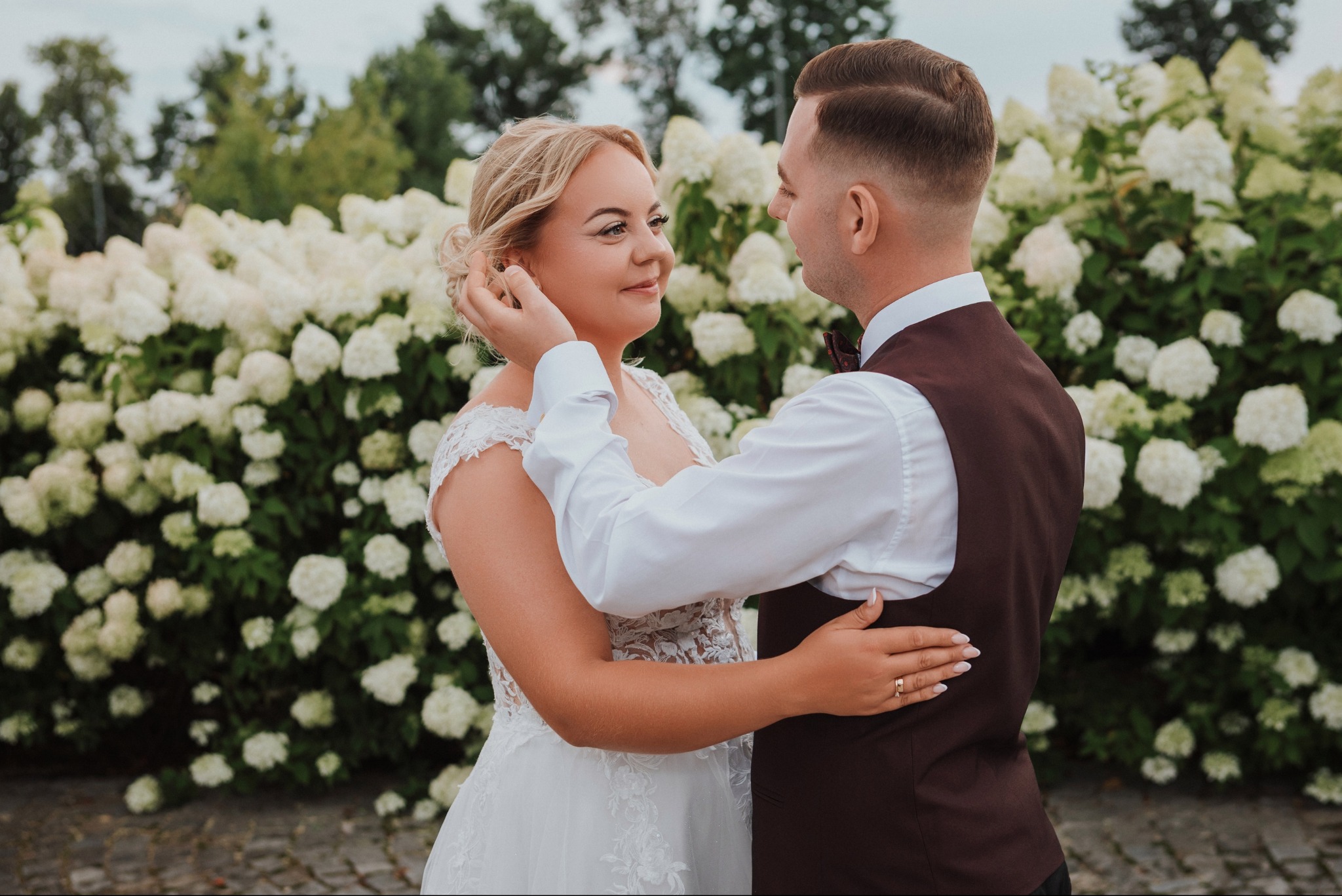 Uśmiechnięta panna młoda w koronkowej sukni, dotykana przez pana młodego w brązowej kamizelce, na tle kwitnących hortensji.