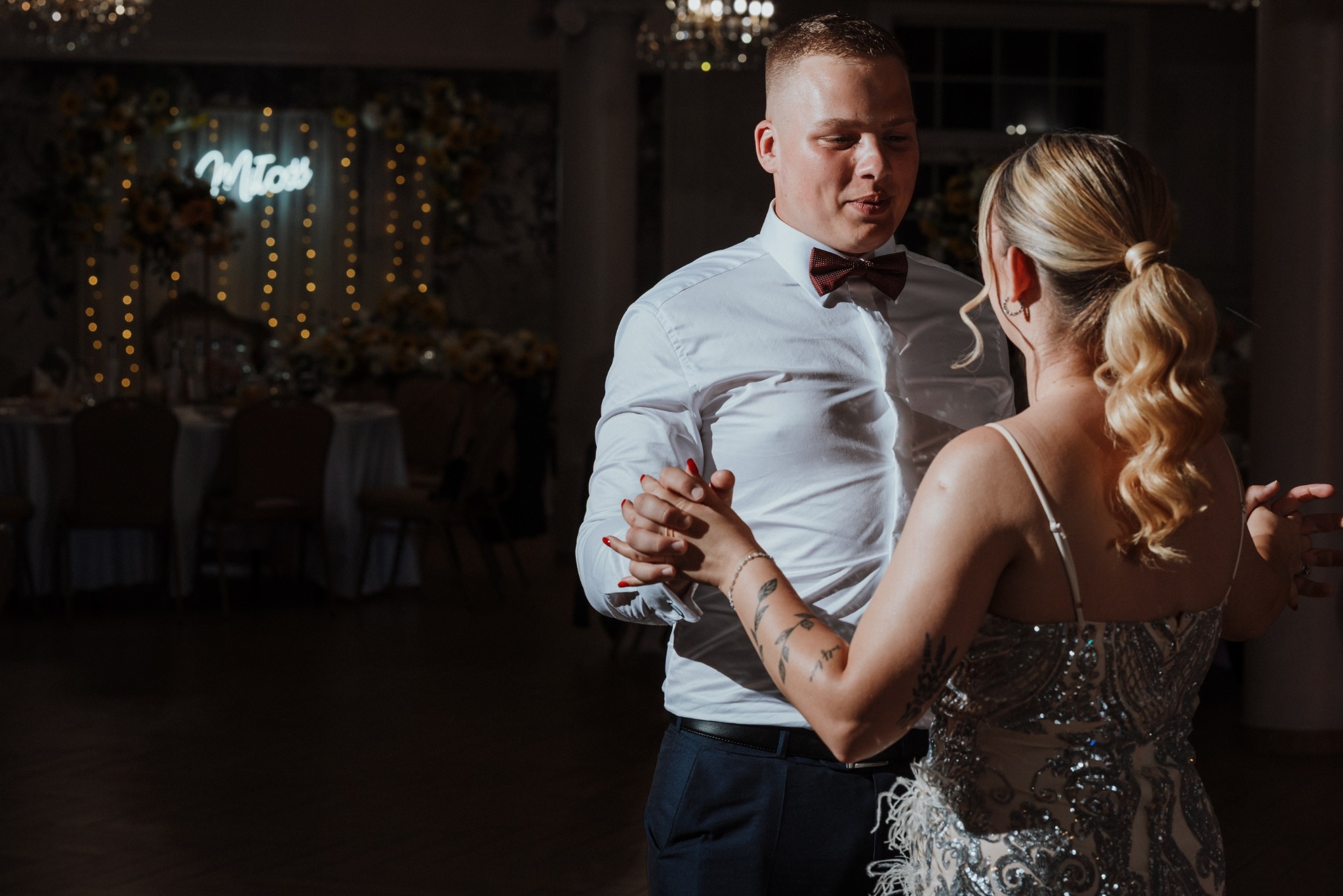 Para młoda tańczy na weselu, on w białej koszuli i bordowej muszce, ona w sukni z cekinami. W tle dekoracja kwiatowa i neonowy napis.