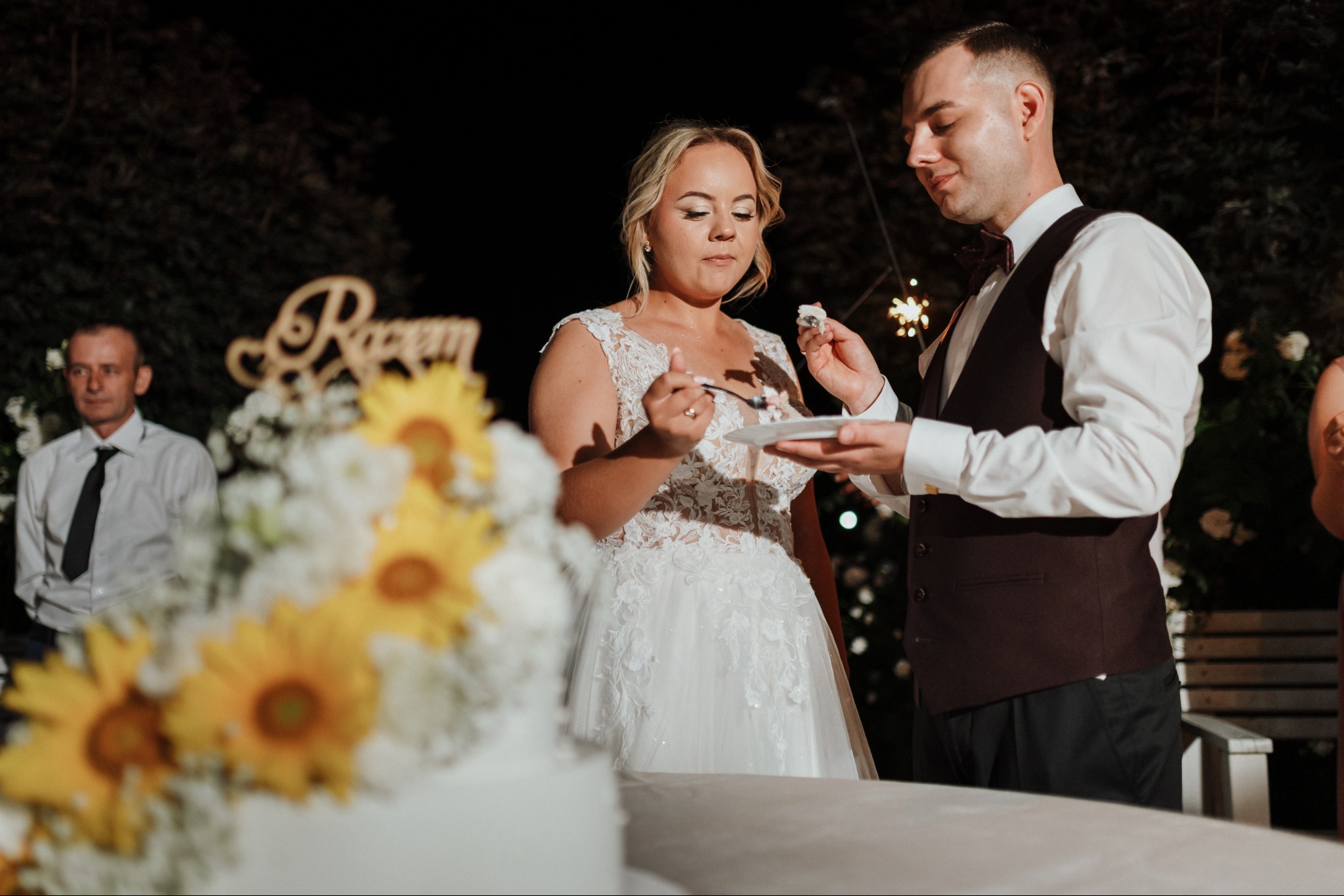 Uśmiechnięta para młoda kroi tort weselny, dekorowany słonecznikami. W tle gość weselny i dekoracja z napisem. Ciepłe światło i radosna atmosfera.