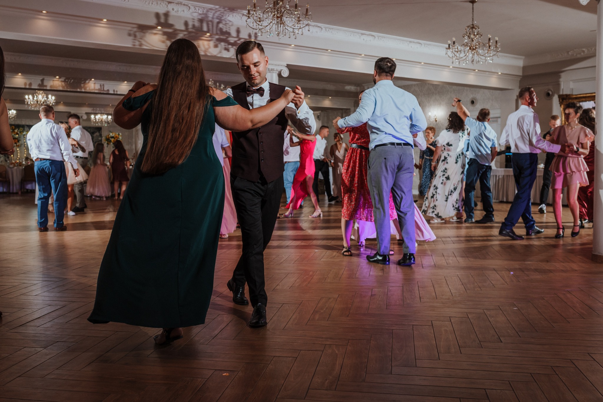 Para tańczy na parkiecie podczas uroczystości. W tle widoczni goście i dekoracje sali balowej z żyrandolami. Mężczyzna w muszce i kamizelce prowadzi partnerkę w zielonej sukni.
