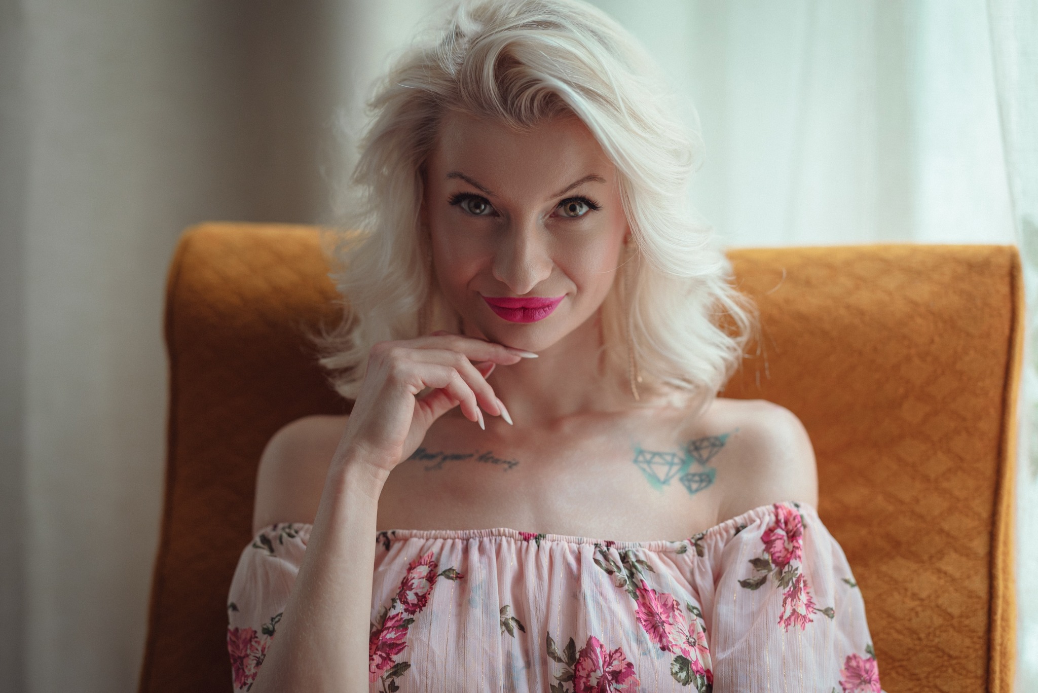 Portret kobiety z platynowymi włosami, w kwiecistej bluzce, siedzącej w fotelu. Widoczny tatuaż na ramieniu i intensywny makijaż ust. Ujęcie z bliska, miękkie światło.