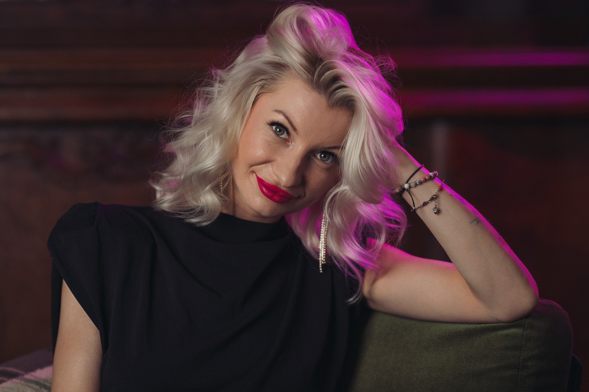Portret kobiety z blond włosami, czerwonymi ustami i czarną bluzką, opierającej się na zielonym fotelu. W tle ciemne drewno i fioletowe światło.