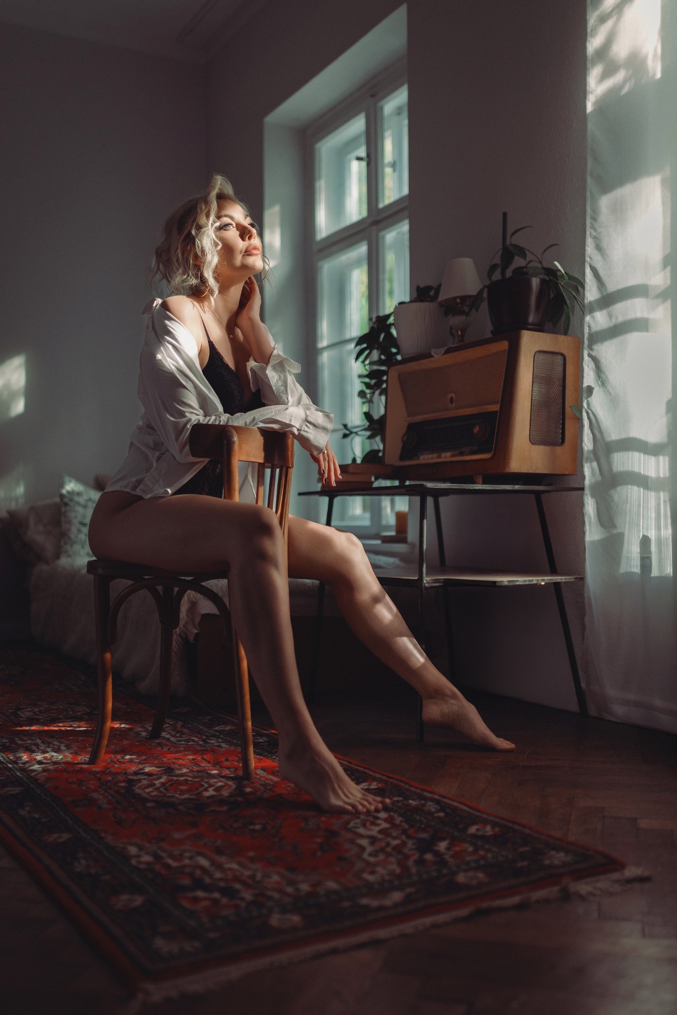 Kobieta w bieliźnie i koszuli siedzi na krześle w pokoju z dywanem i radiem, spoglądając w górę na światło wpadające przez okno. Klimatyczne, intymne ujęcie.