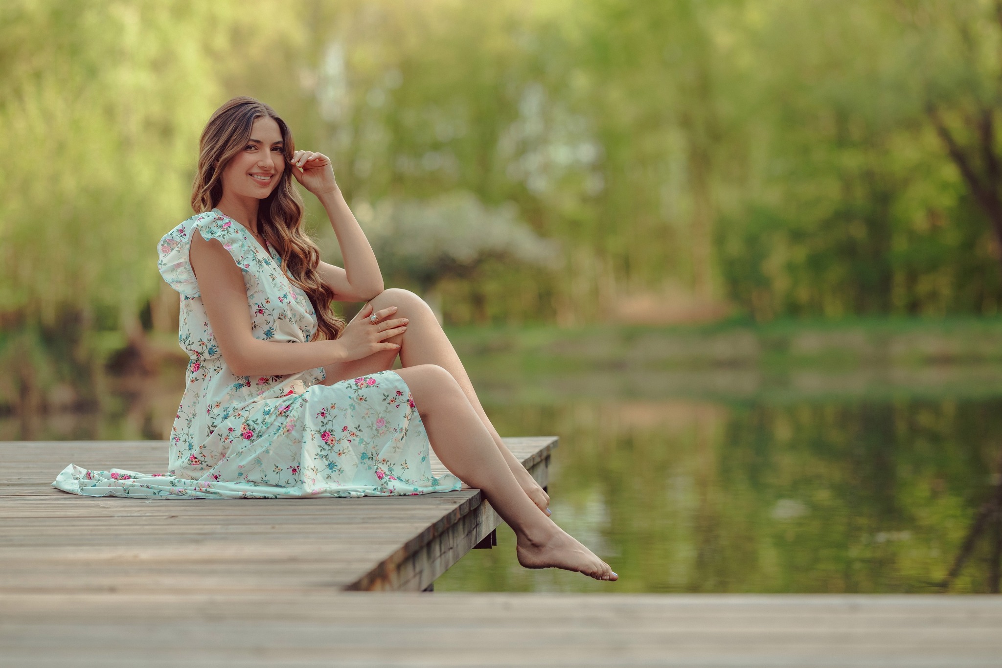 Uśmiechnięta brunetka w kwiecistej sukience siedzi boso na drewnianym pomoście nad wodą, z zalesionym brzegiem w tle. Delikatne, naturalne światło.