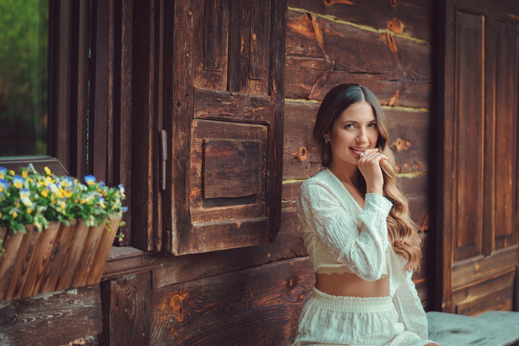 Uśmiechnięta kobieta w białym stroju pozuje na tle drewnianej ściany z oknem i kwiatami w doniczce. Naturalne światło, ciepła kolorystyka, swobodna atmosfera.