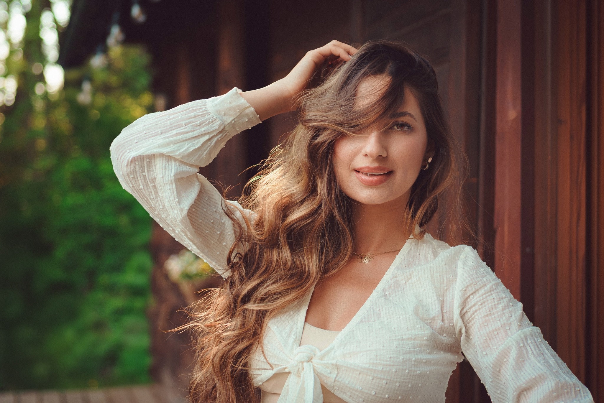 Portret uśmiechniętej brunetki z długimi, falowanymi włosami, w białej bluzce z długim rękawem, pozującej na tle drewnianej ściany i zieleni.