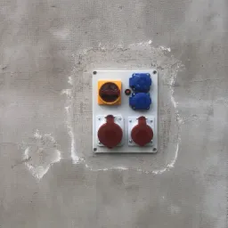 Panel elektryczny z wyłącznikiem i gniazdami przemysłowymi w kolorach czerwonym i niebieskim, zamontowany na surowej, szarej ścianie z widocznymi śladami po tynku.