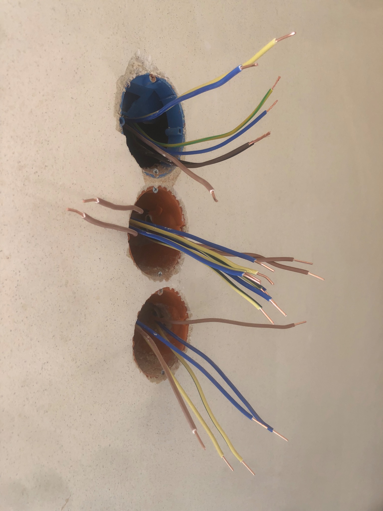 Trzy puszki elektryczne w ścianie z wystającymi przewodami w kolorach: niebieskim, żółtym, zielonym i brązowym, przygotowane do podłączenia.